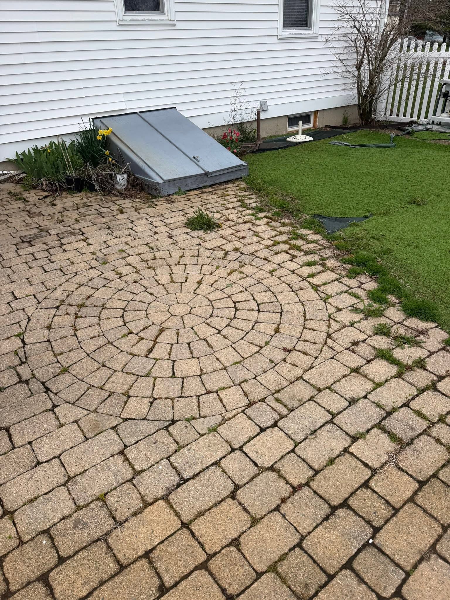 Brick paver patio beside white house with a small metal cellar door and grassy yard.