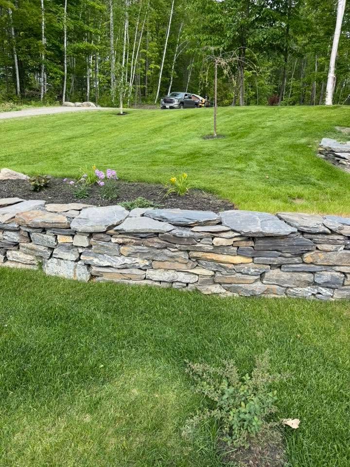 Stone retaining wall beside a green lawn with trees and a parked car in the background