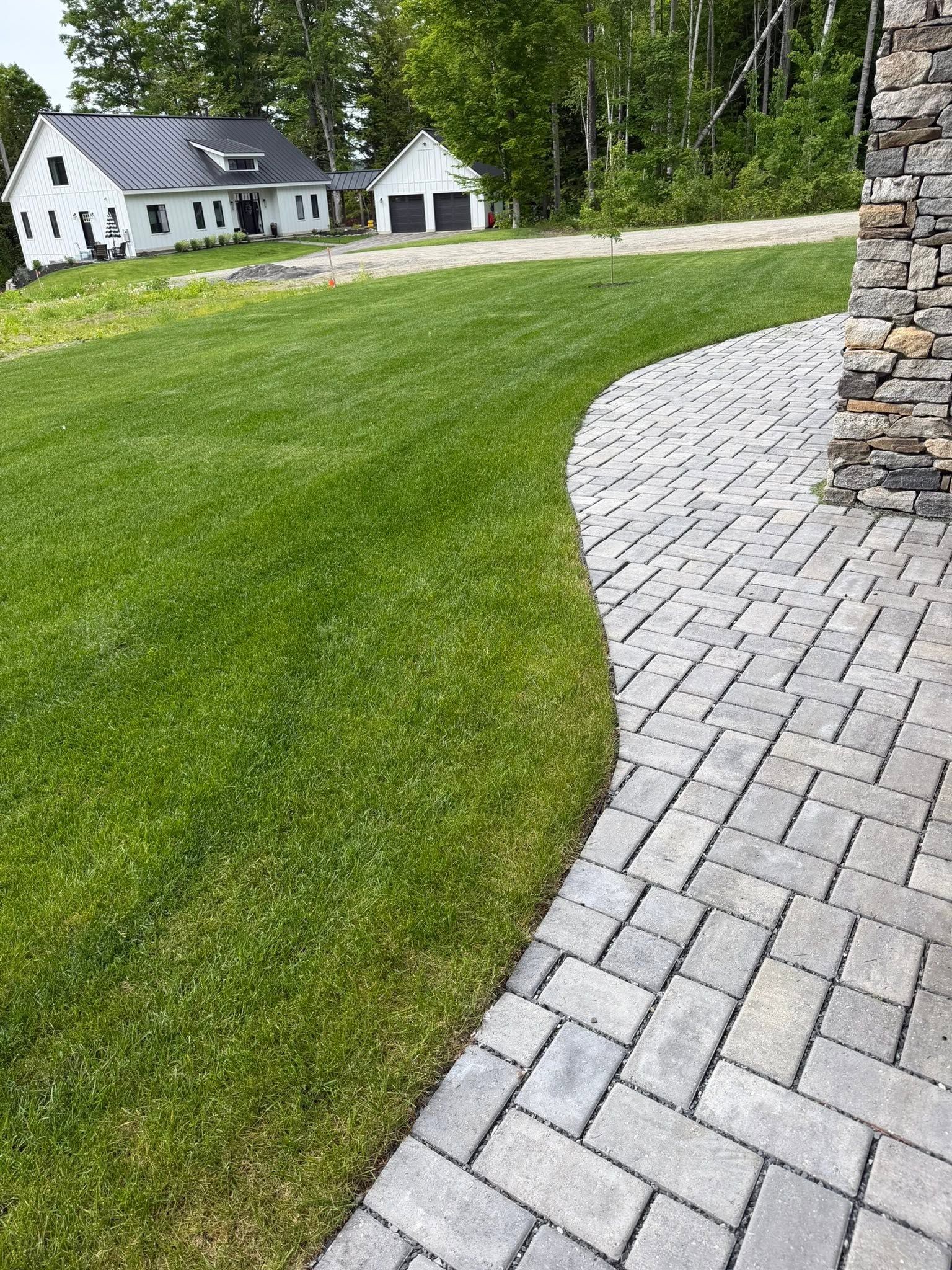 Paved driveway beside a green lawn leading to a white house and trees.
