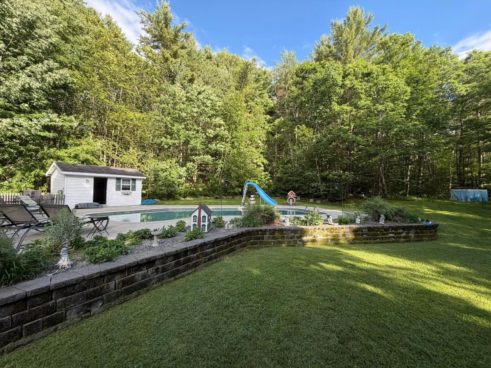 Backyard pool with lounge chairs, slide, and small shed beside a grassy lawn and wooded trees