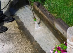 A green toy sprinkler sprays water on a concrete path beside a wooden garden border.