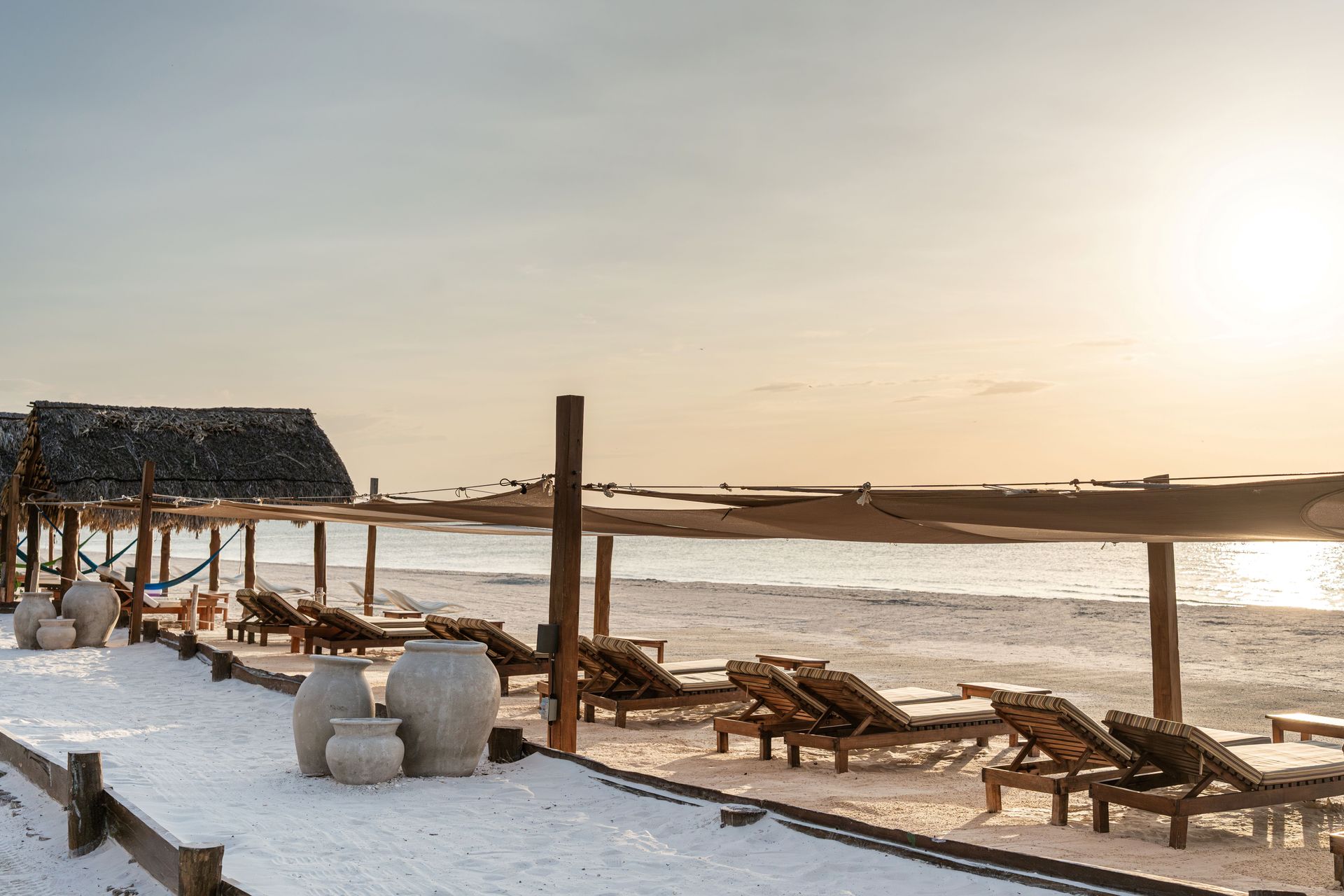 Beach scene with snow-covered sand, wooden lounge chairs, thatched roof shelter, and setting sun.