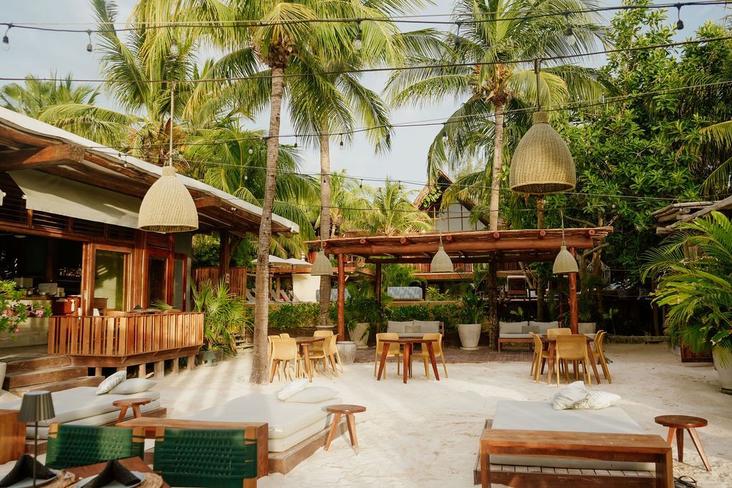 Outdoor dining area with sand floor, wooden structures, palm trees, and hanging woven lamps.
