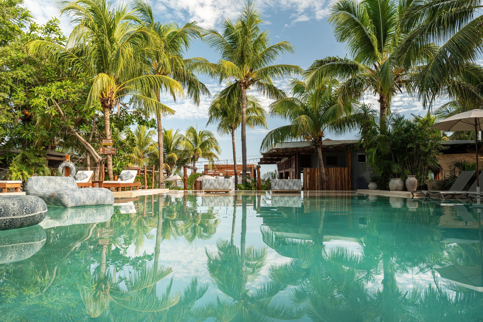 Tropical pool with palm trees reflected in the turquoise water. A wooden bar and seating area are in the background.