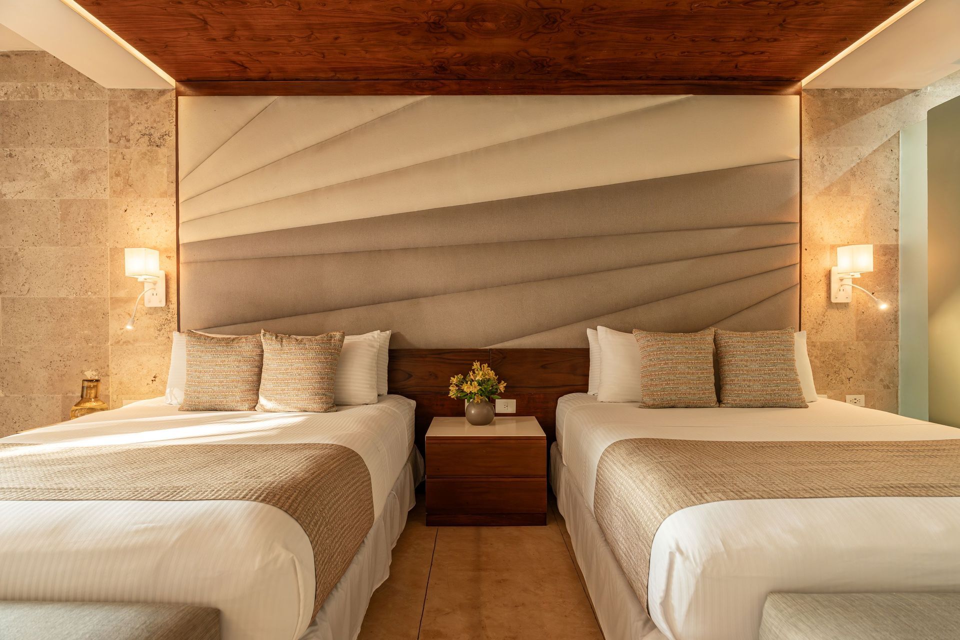 Two beds with cream and gold bedding, nightstand, and beige geometric headboard in a hotel room.