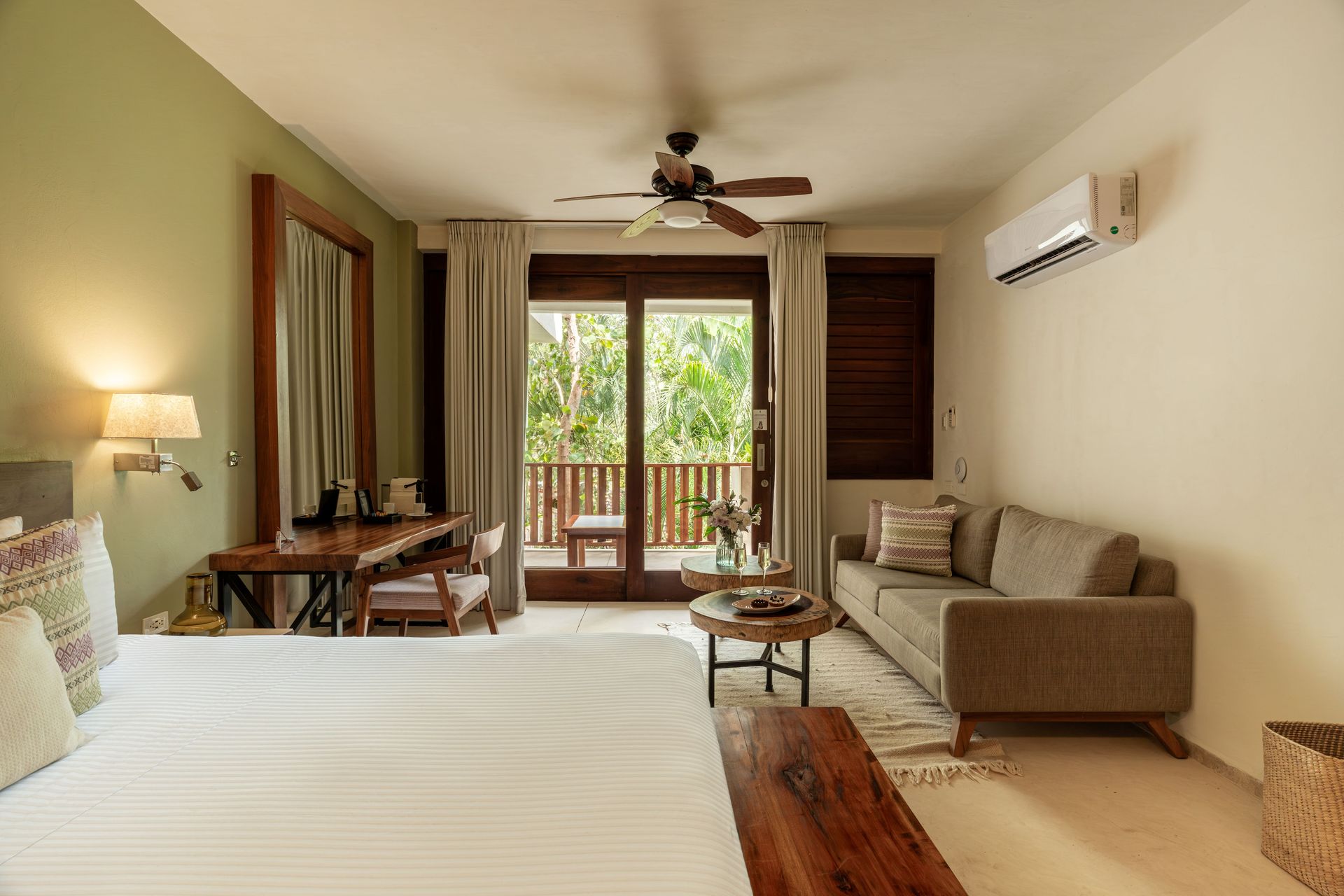 Bedroom with balcony: bed, sofa, desk, open doorway to a balcony with green foliage, light colors.