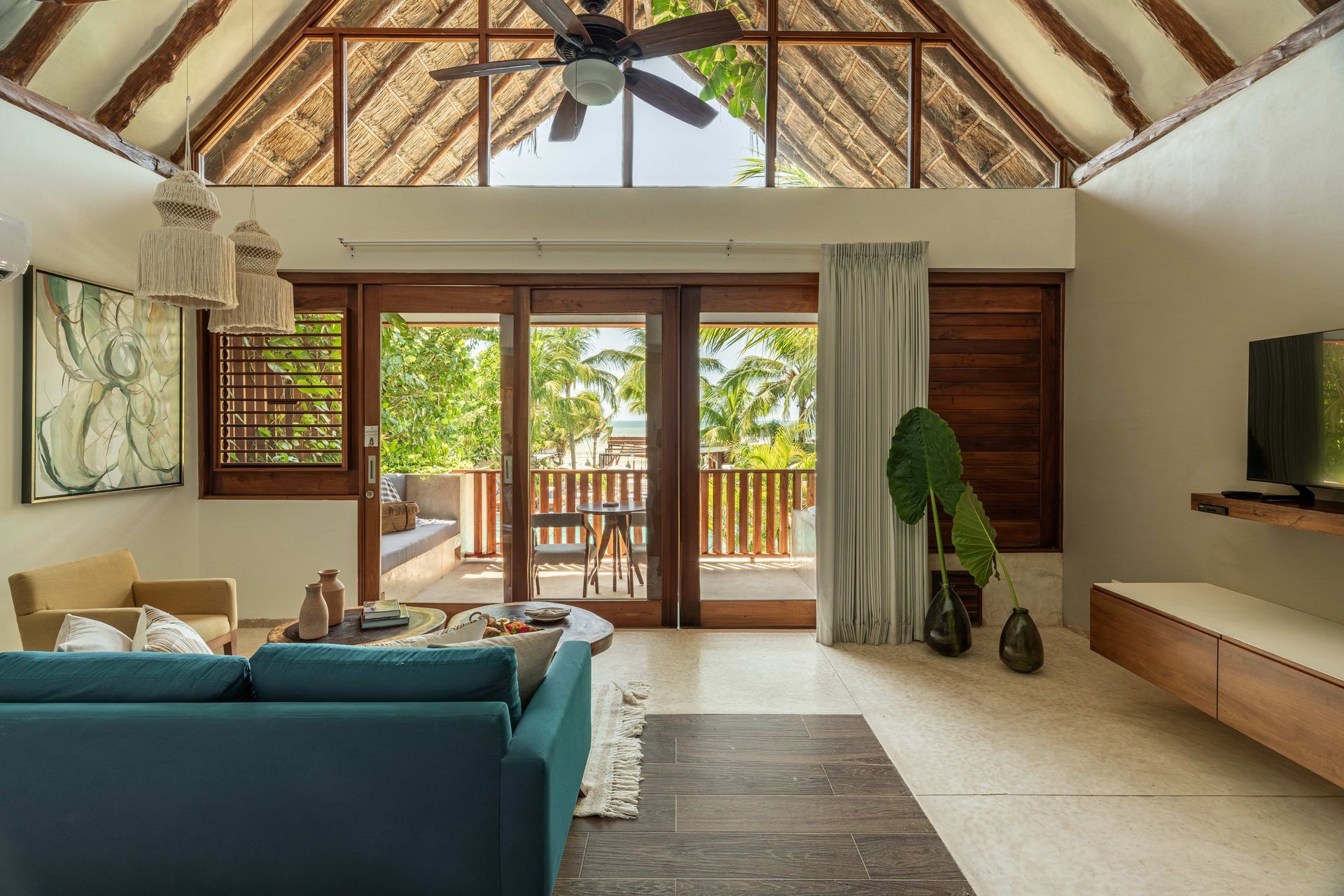 Living room with teal sofa, wooden balcony doors, and exposed thatched roof.