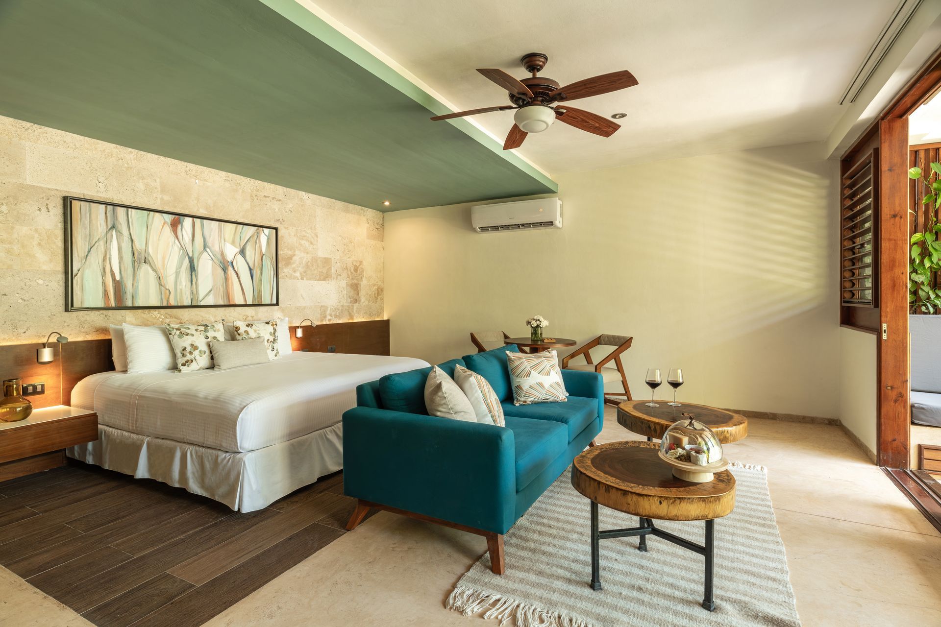Cozy hotel room with king bed, teal sofa, small round tables, and an outdoor balcony.