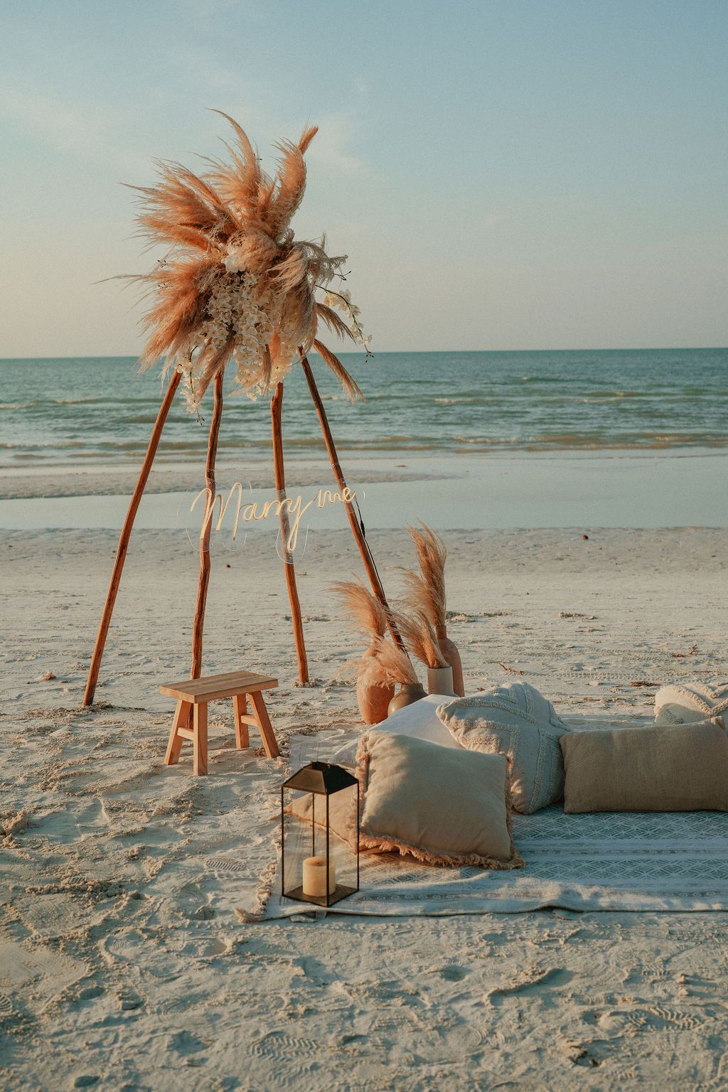 Beach scene with teepee-like structure decorated with pampas grass. Pillows and a lantern sit nearby.