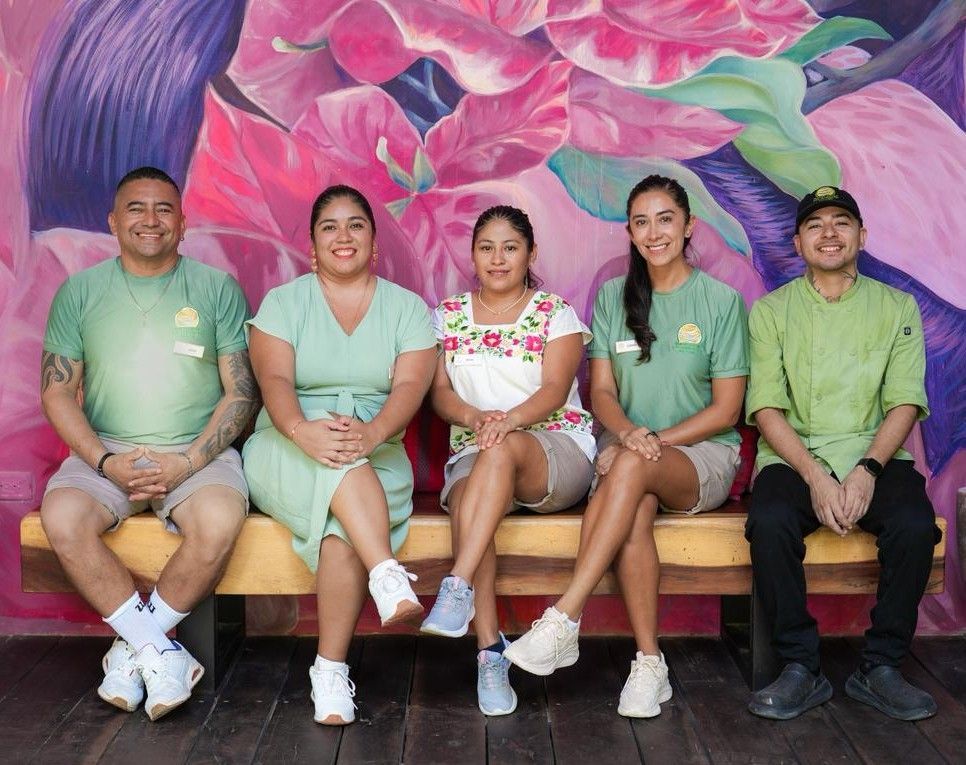 Five people in green shirts sit on a bench in front of a colorful floral mural.