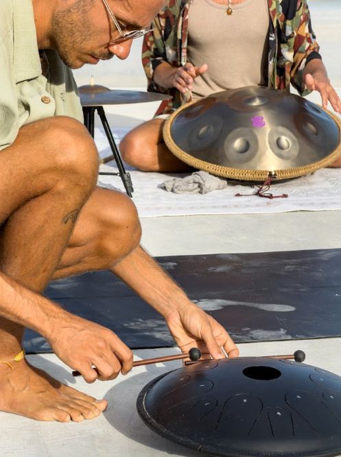 Two people tuning handpans on a white cloth, one hammering a black steel drum on the floor