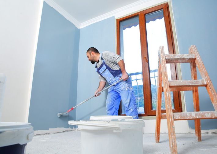 Painter roller-coating a wall in a bright room with a wooden ladder nearby