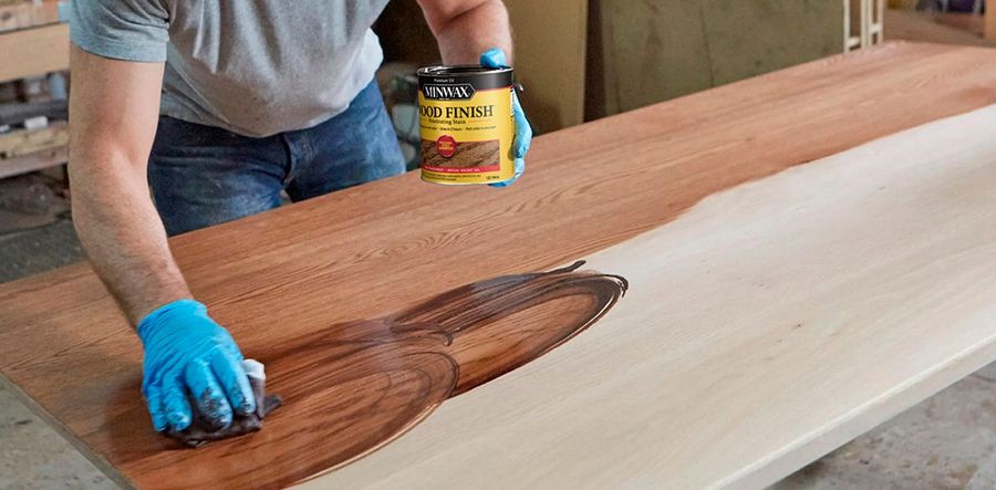 Person applying wood stain to a plank on a workbench, with a blue-gloved hand and can of stain.