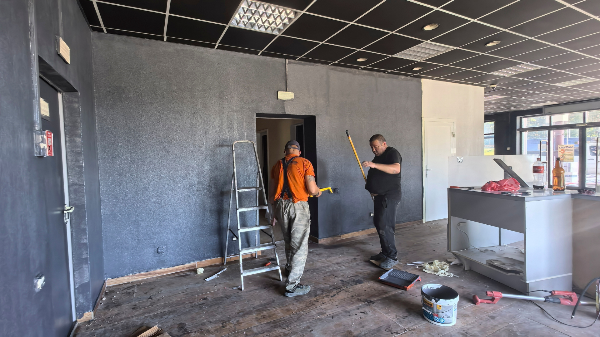 Two workers painting a dark wall in a room with ladders, tools, and renovation debris