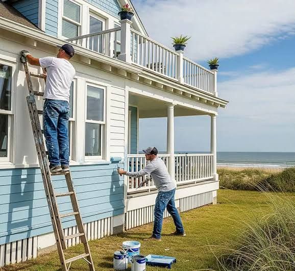 Two painters working on the exterior of a seaside house, one on a ladder and one near the porch.