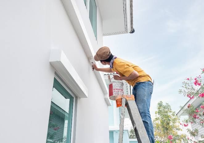 Person painting a building exterior from a ladder, holding a paint bucket.