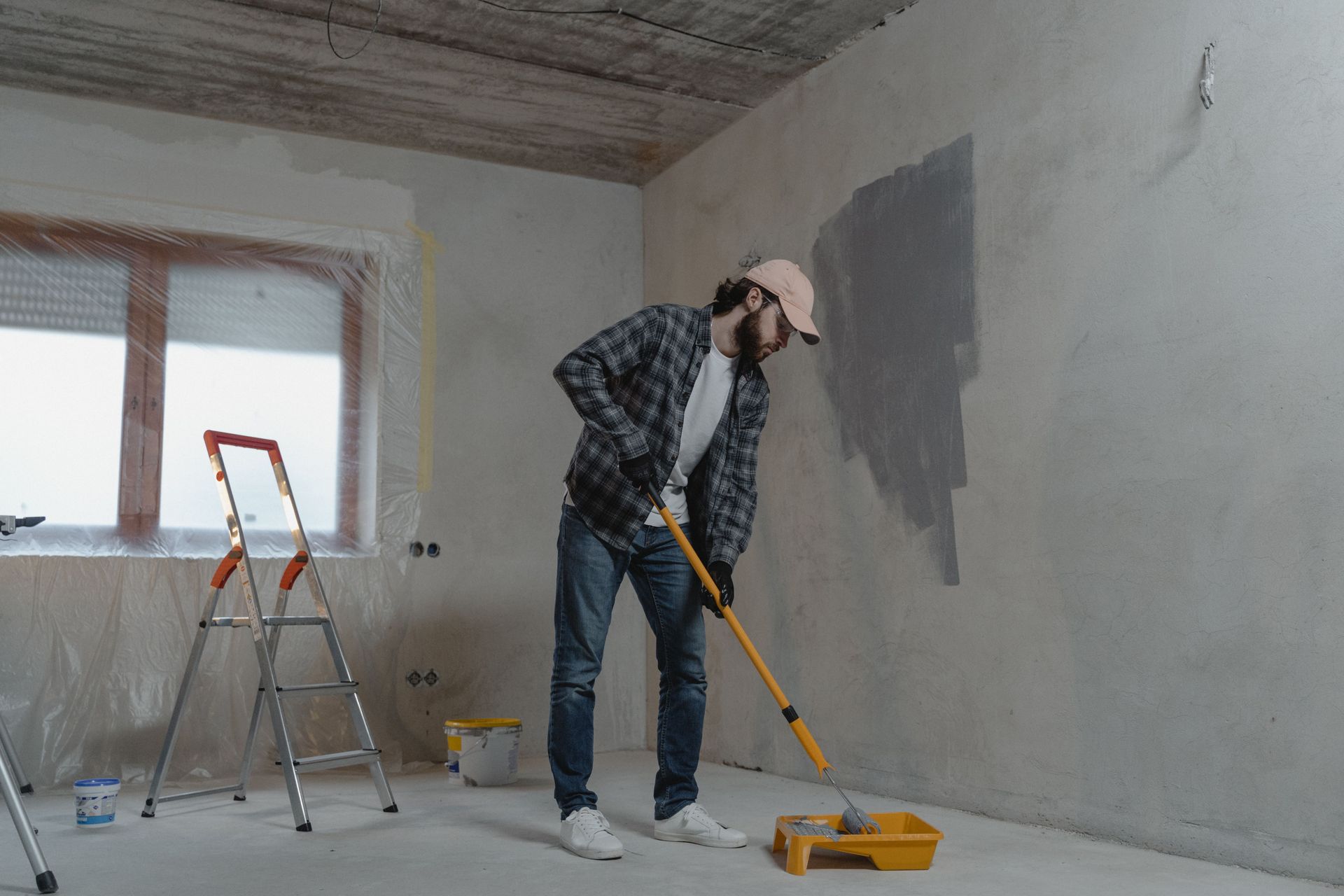 Person painting an interior wall with a roller in a bare, unfinished room.