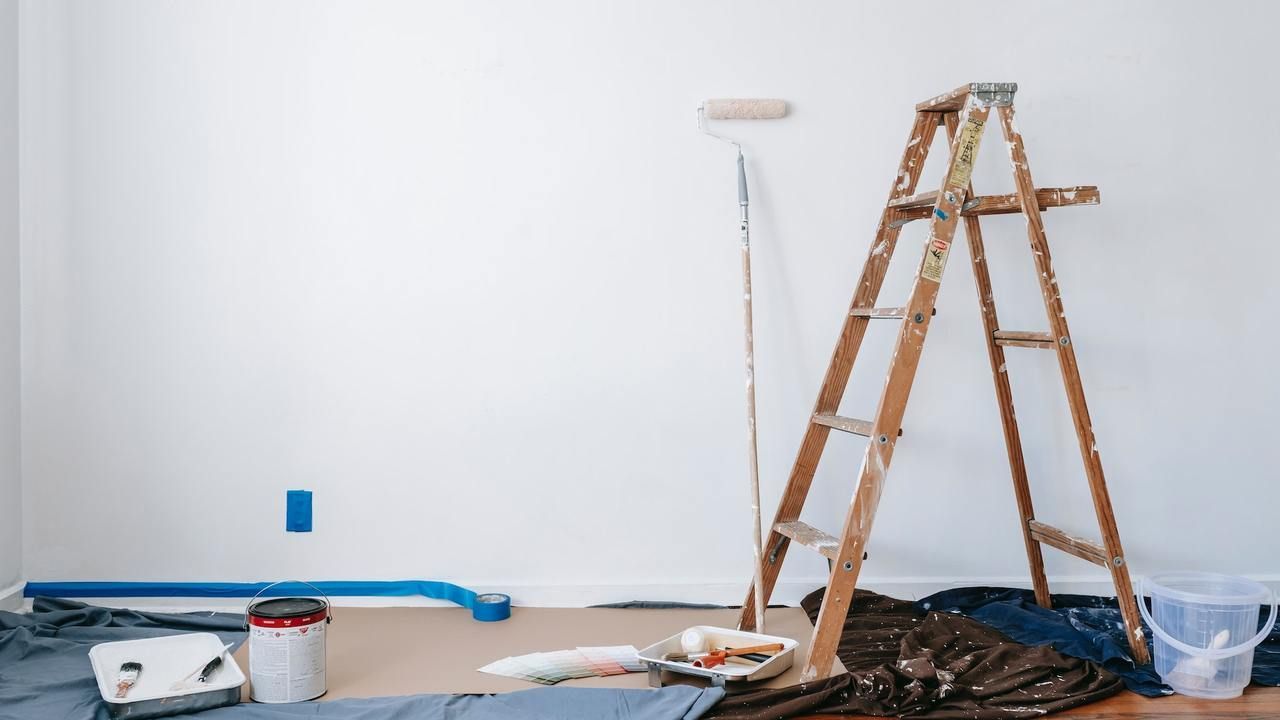 Painted white wall with ladder, paint roller, buckets, tarp, and tools on the floor in a work area.