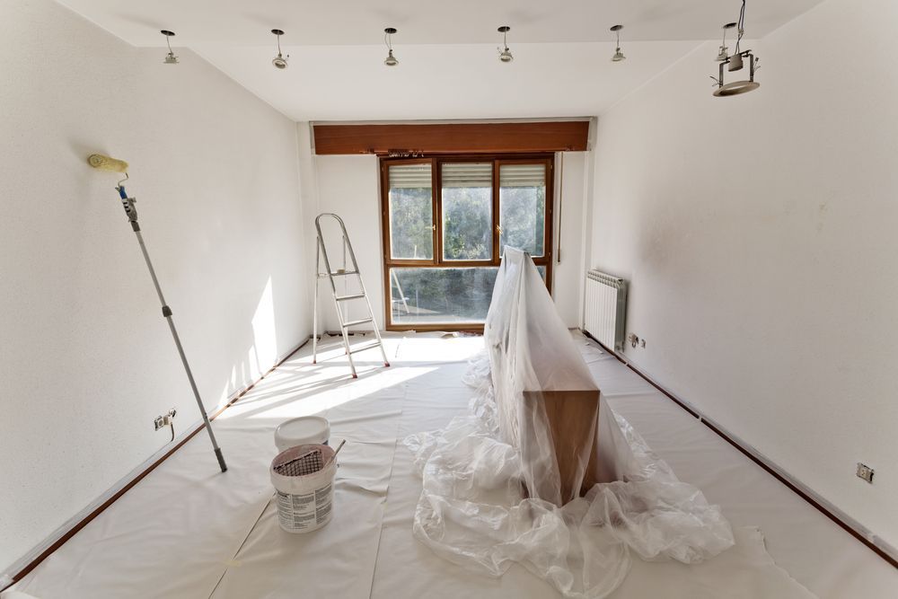 Empty white room under renovation with plastic-covered furniture, paint bucket, ladder, and window light