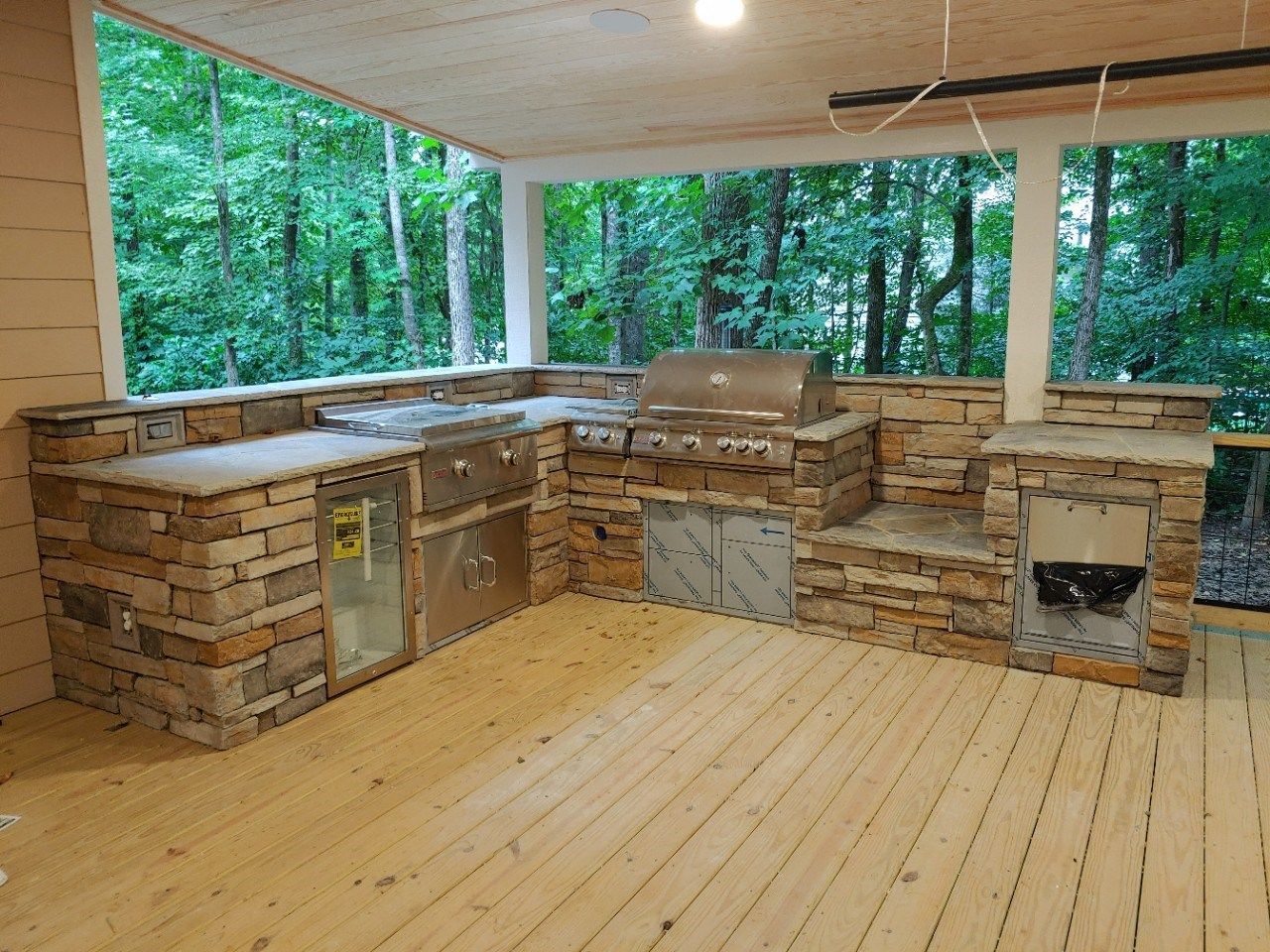 Outdoor kitchen with stone counters, stainless steel grill, and appliances on a wooden deck overlooking a forest.