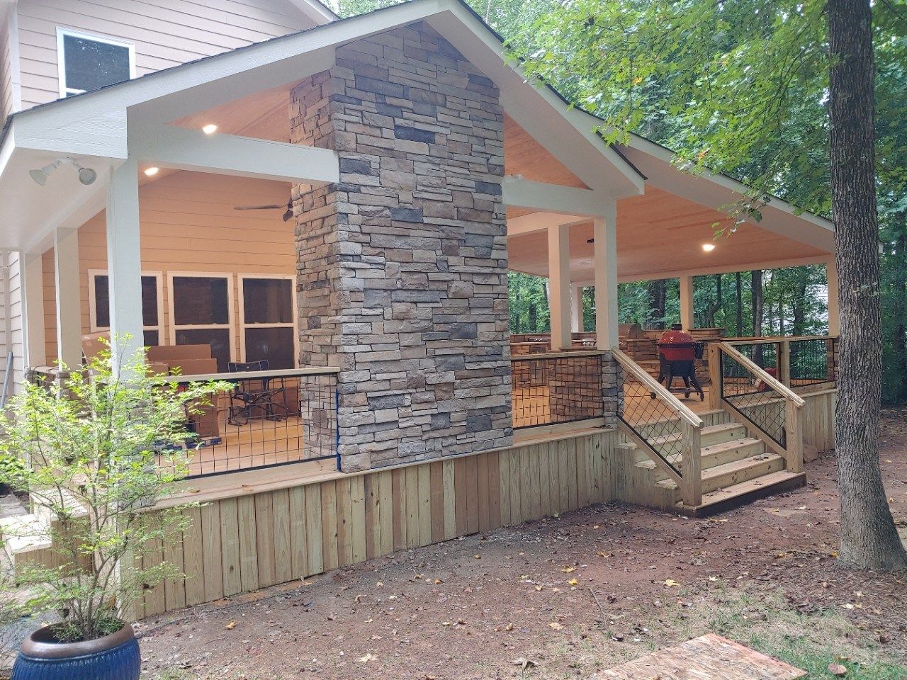 A spacious, covered outdoor deck with a stone fireplace, wood railings, and stairs, set against a wooded background.