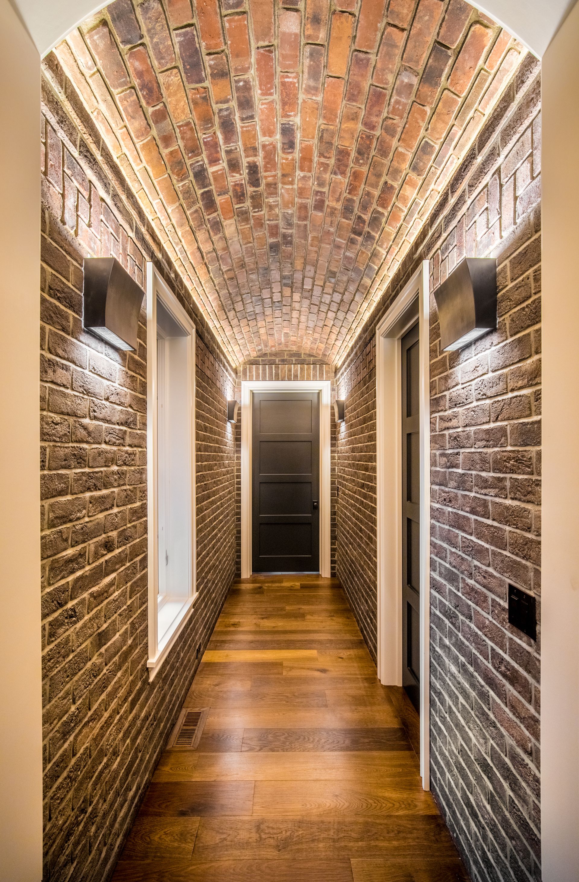 Long, brick hallway with arched ceiling, sconce lights, and dark wood floor.