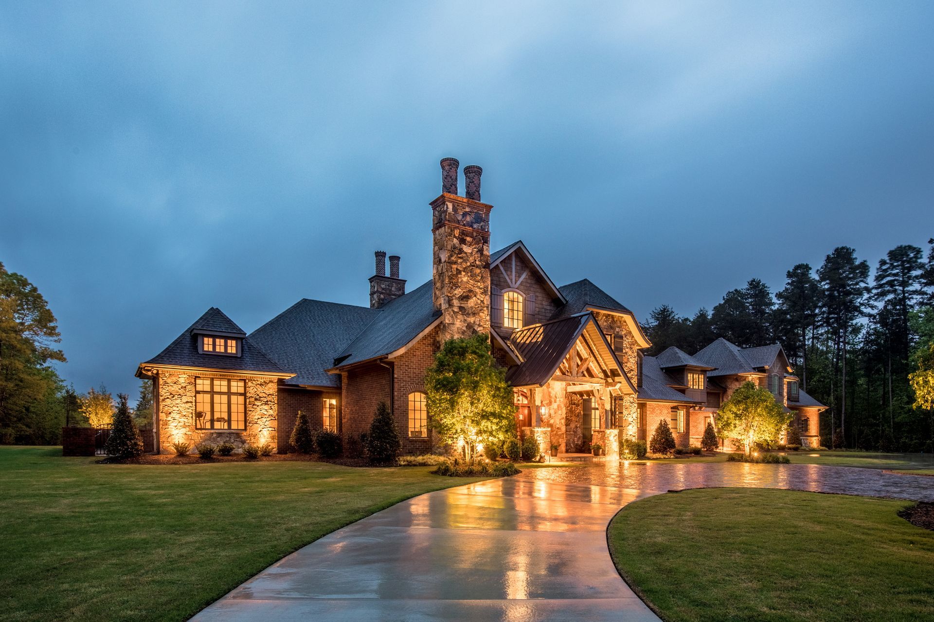 Stone mansion illuminated at dusk with a curved driveway, surrounded by lawn and trees.