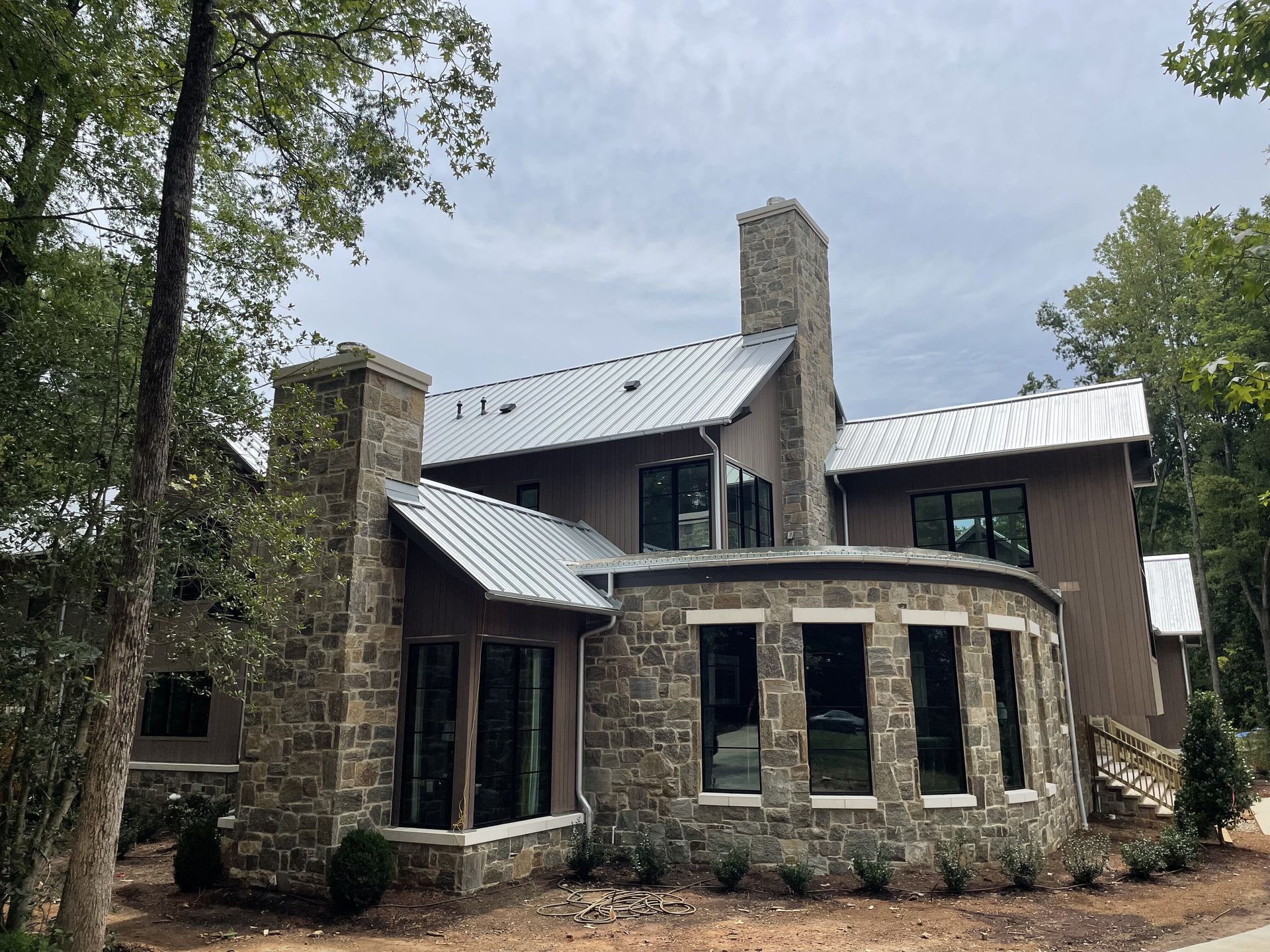 Stone and stucco house with metal roof, chimney, and large windows set amidst trees under a cloudy sky.