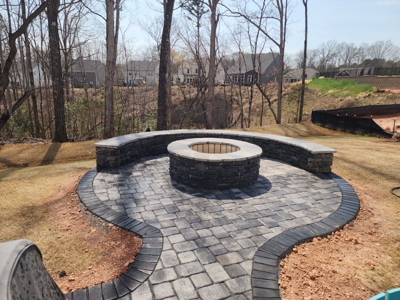 A circular stone fire pit sits in the center of a paved patio with a curved stone bench, set in a backyard near woods.