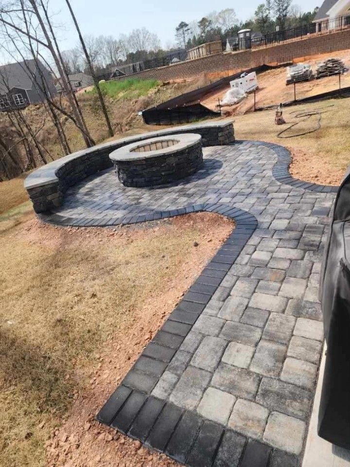 A stone patio featuring a circular fire pit and curved seating wall, surrounded by a lawn in a residential backyard.