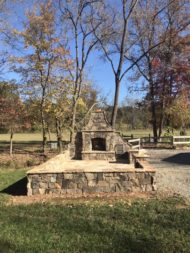 An outdoor stone fireplace and patio area set in a grassy yard with trees under a clear blue sky.