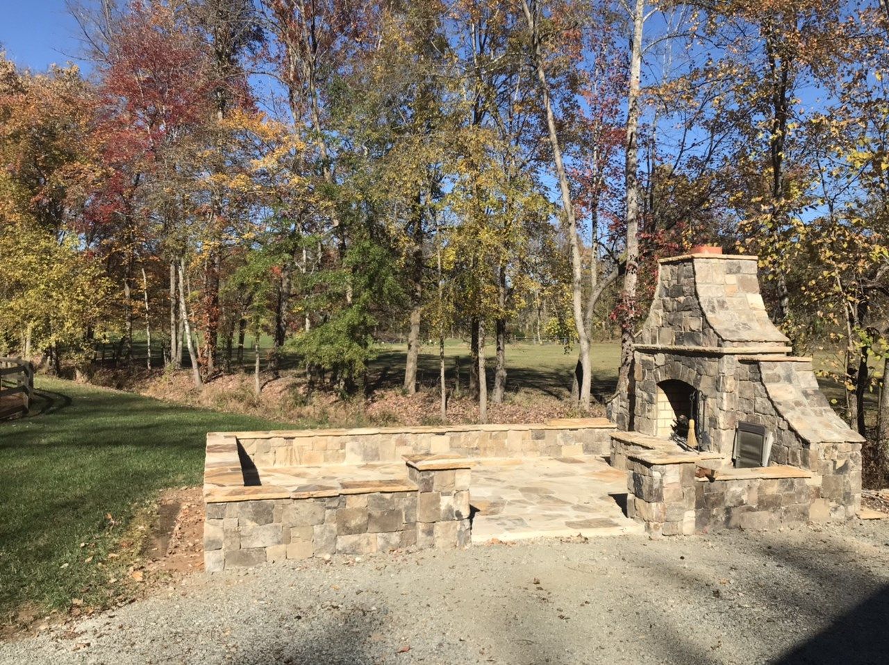 An outdoor stone fireplace and patio with built-in stone seating, situated on a gravel surface near a wooded area.