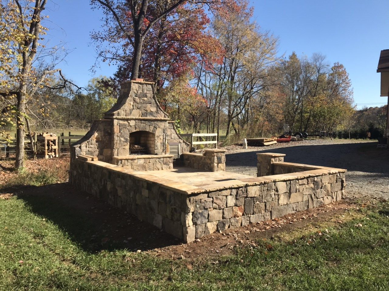 An outdoor stone patio with a built-in fireplace and low perimeter walls, set in a grassy yard under a clear blue sky.