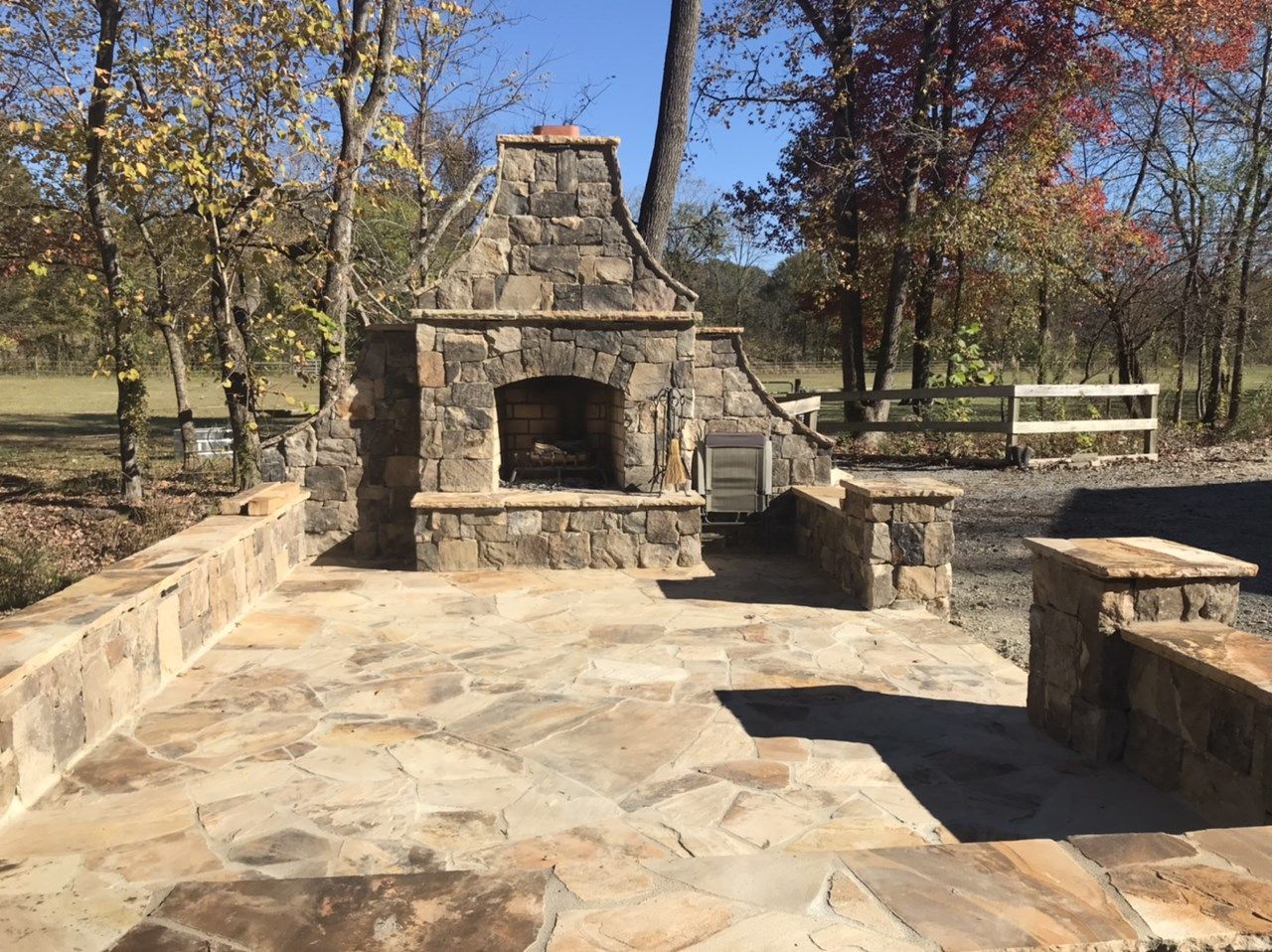 An outdoor stone patio featuring a built-in stone fireplace and seating walls on a sunny day with trees in the background.