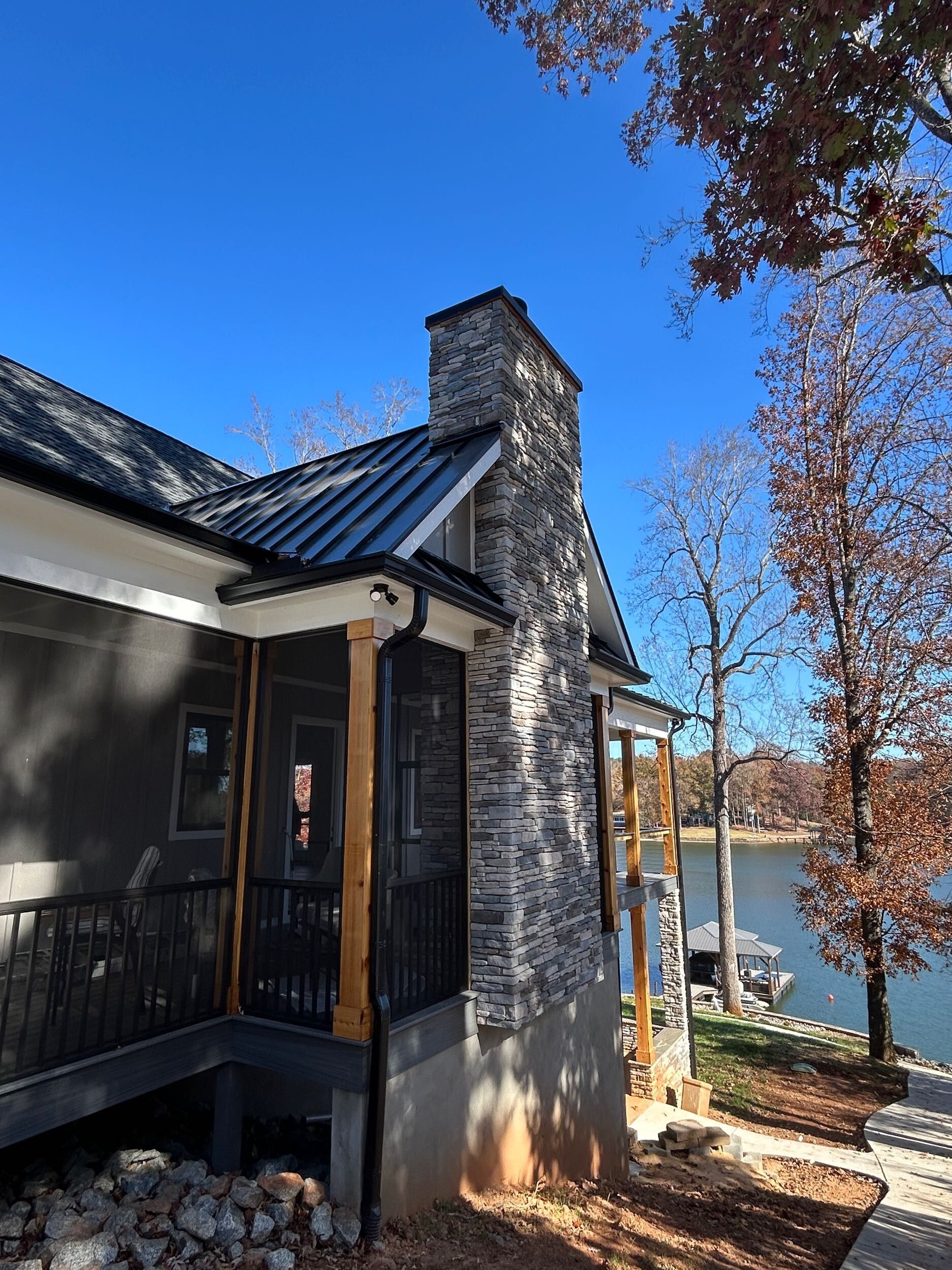 A stone fireplace chimney stands on the side of a modern house overlooking a lake on a sunny day.