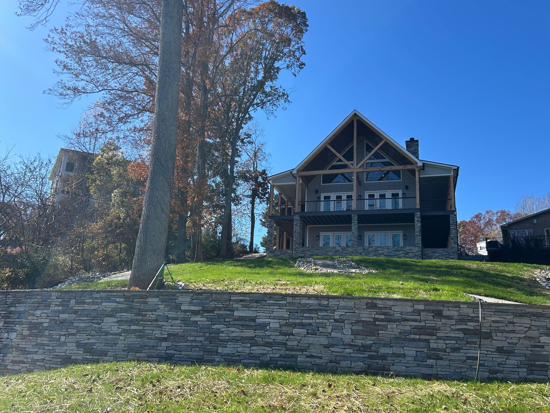 A large, modern, multi-story wooden house with a stone base sits atop a grassy hill behind a tall stacked stone wall.