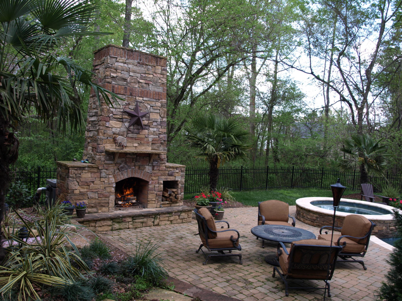 Outdoor fireplace with brick and stone, patio furniture, and a hot tub in a natural setting.