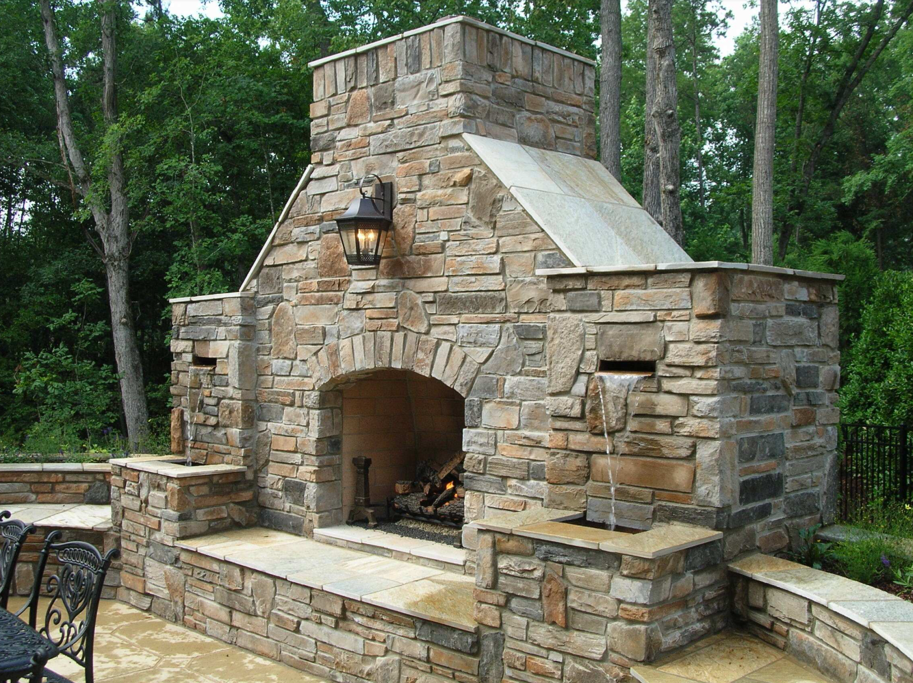 Stone outdoor fireplace with a burning fire, surrounded by greenery.
