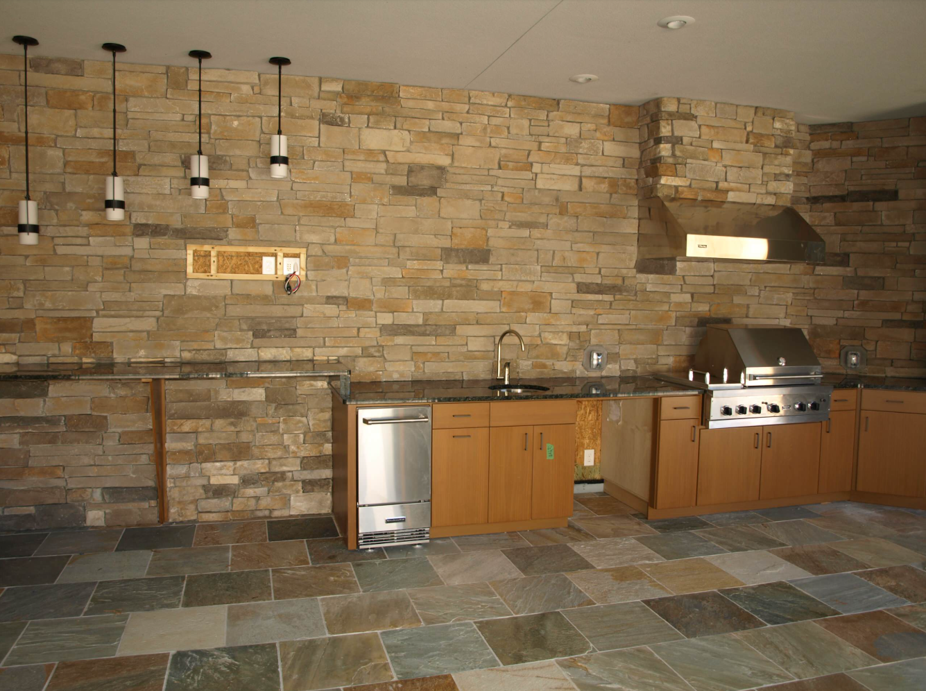 Outdoor kitchen with stone wall, stainless steel appliances, and wood cabinets.