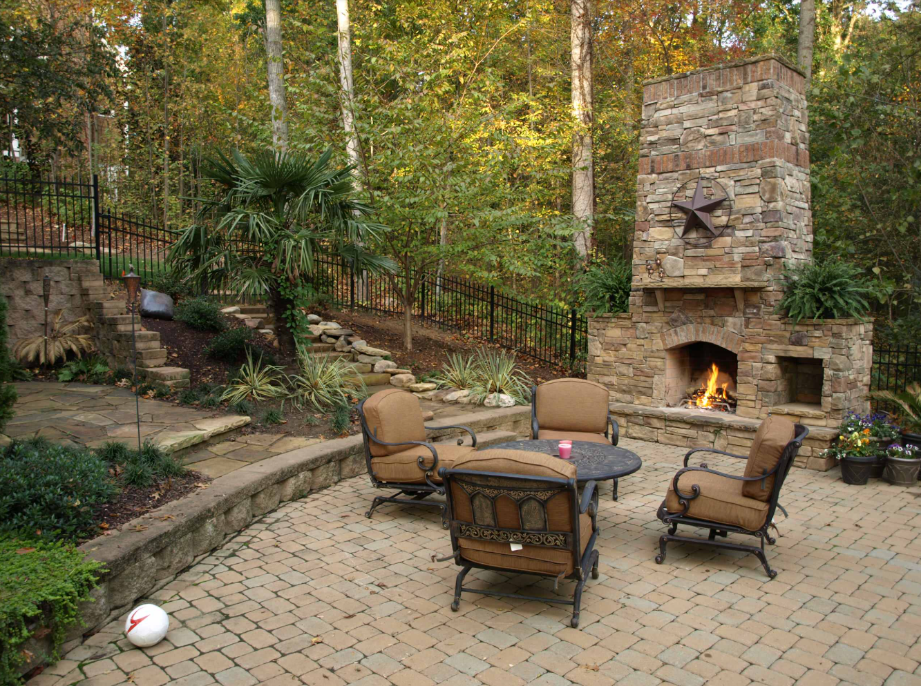 Outdoor patio with fireplace, seating, and lush greenery. Stone fireplace with a fire, brick patio.