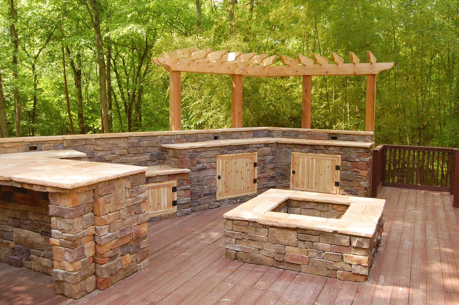 Outdoor kitchen with stone counters, wooden pergola, and fire pit on a wooden deck. Lush green trees in background.