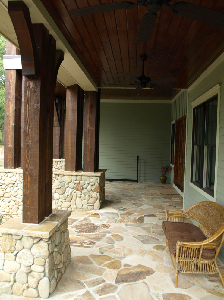 Covered porch with stone columns, flagstone flooring, wooden ceiling, and wicker bench.