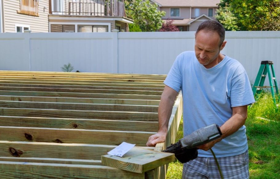 Man installing wooden deck boards in a backyard, using a drill beside a partially built frame.