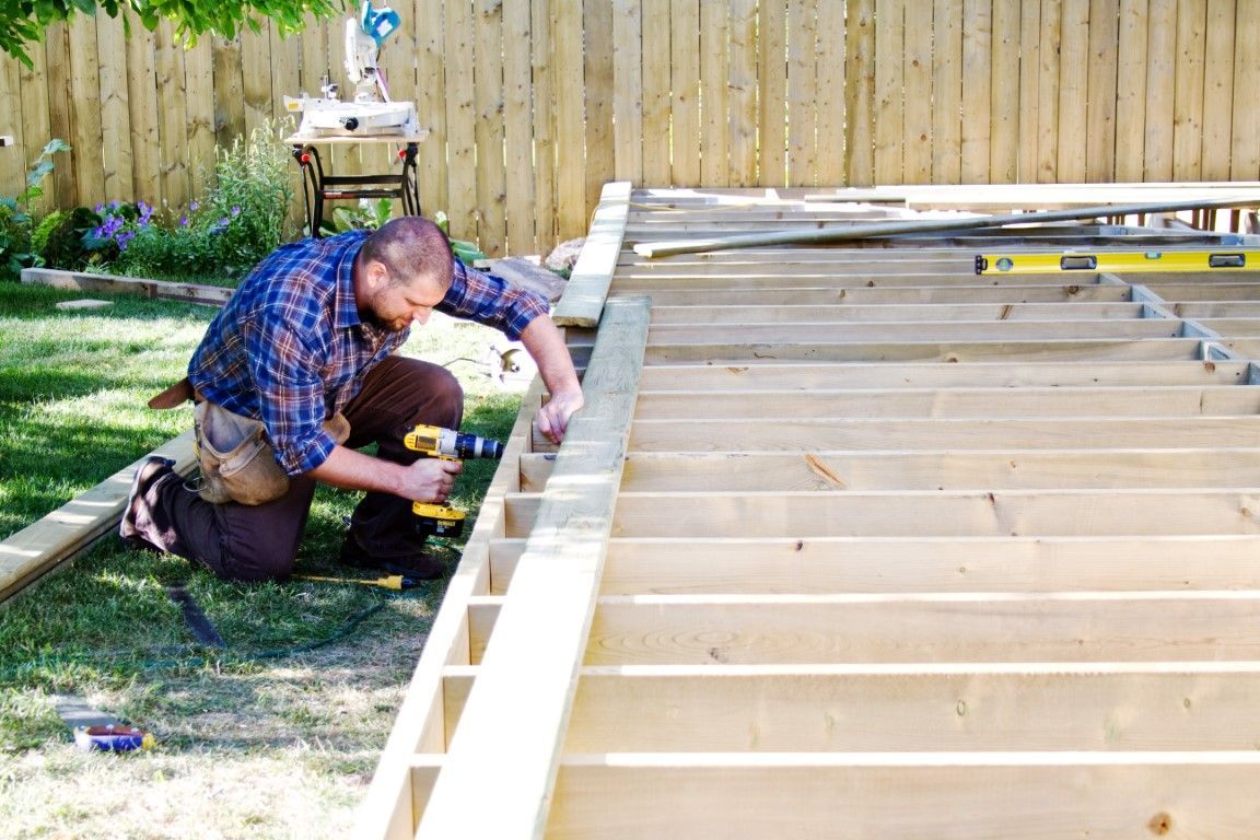 Man kneeling beside a large wooden deck frame, measuring and securing boards in a backyard.