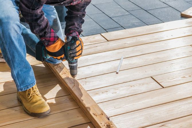 Person drilling a wooden deck board beside a wet surface, wearing jeans and work boots