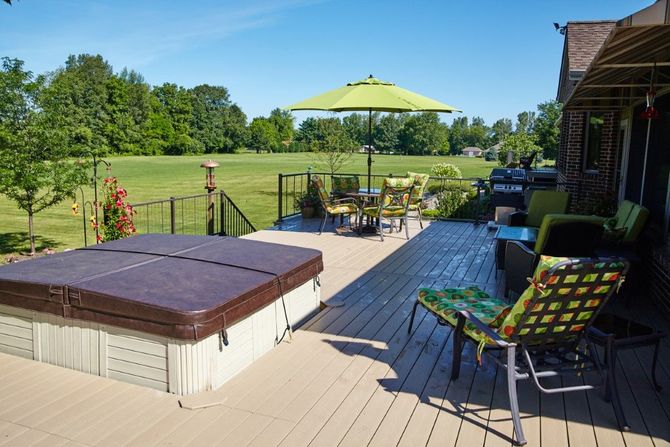 Backyard patio with hot tub, chairs, and green umbrella overlooking a grassy field