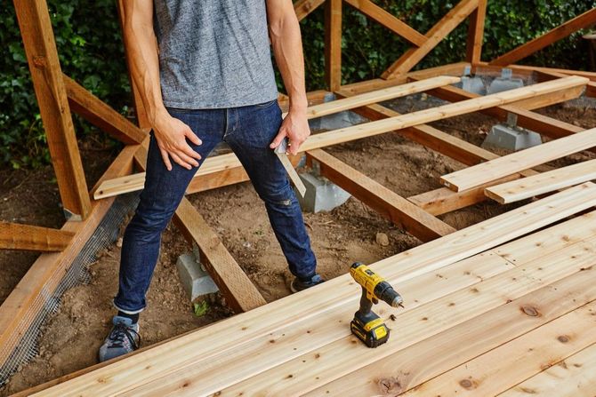 Person standing on a wooden floor frame during house construction, with a yellow drill nearby.