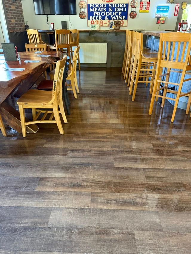 A restaurant interior with wooden tables and chairs set on a brown wood-plank floor, with a bar area in the background.