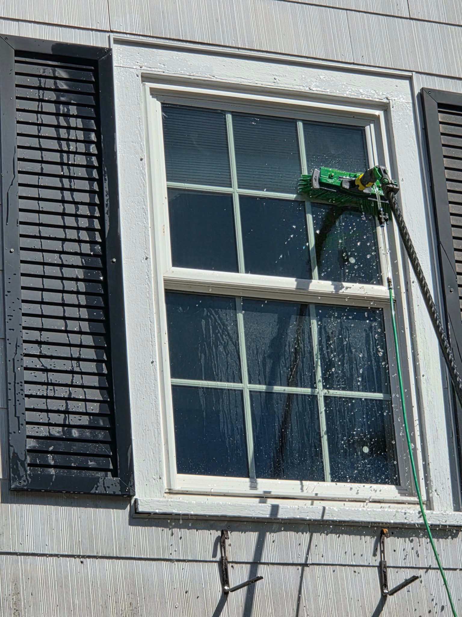 A pole-mounted brush cleans a white-framed window with black shutters, spraying water droplets onto the glass.