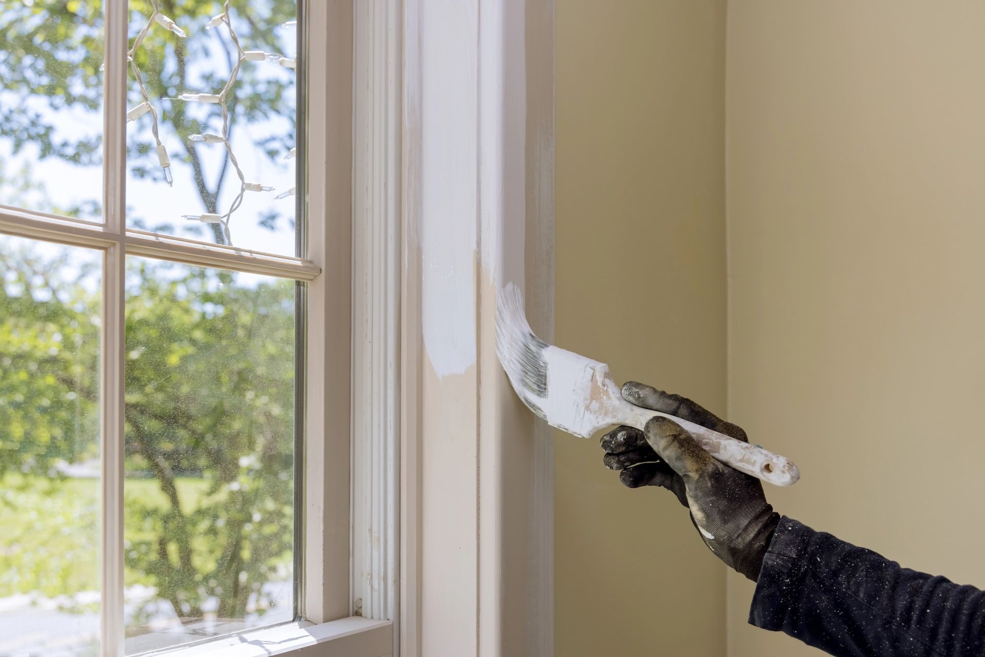 Person in black gloves paints white trim around a window with a brush.